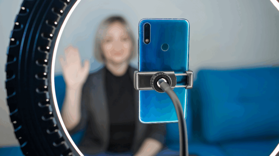 Blurry business woman waving hello while sitting on a blue couch. She is recording herself with a phone connected to a ring light.